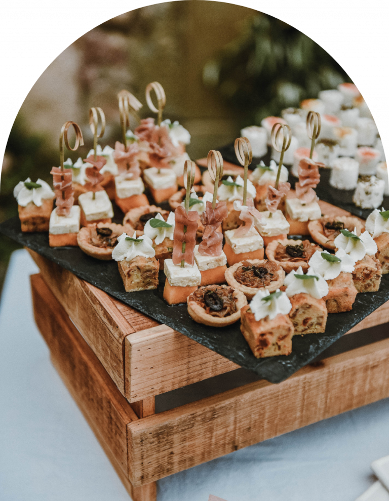 Présentation sur buffet avec mise en volume : petite caisse en bois naturel. Ici, des pièces apéritives dressées sur ardoise pour un effet authentique : tartelettes aux oignons façon pissaladière, mini cakes aux tomates séchées, olives et leur chantilly, gaspacho andalou et brochettes de melon, feta marinée au balsamique et jambon de pays.