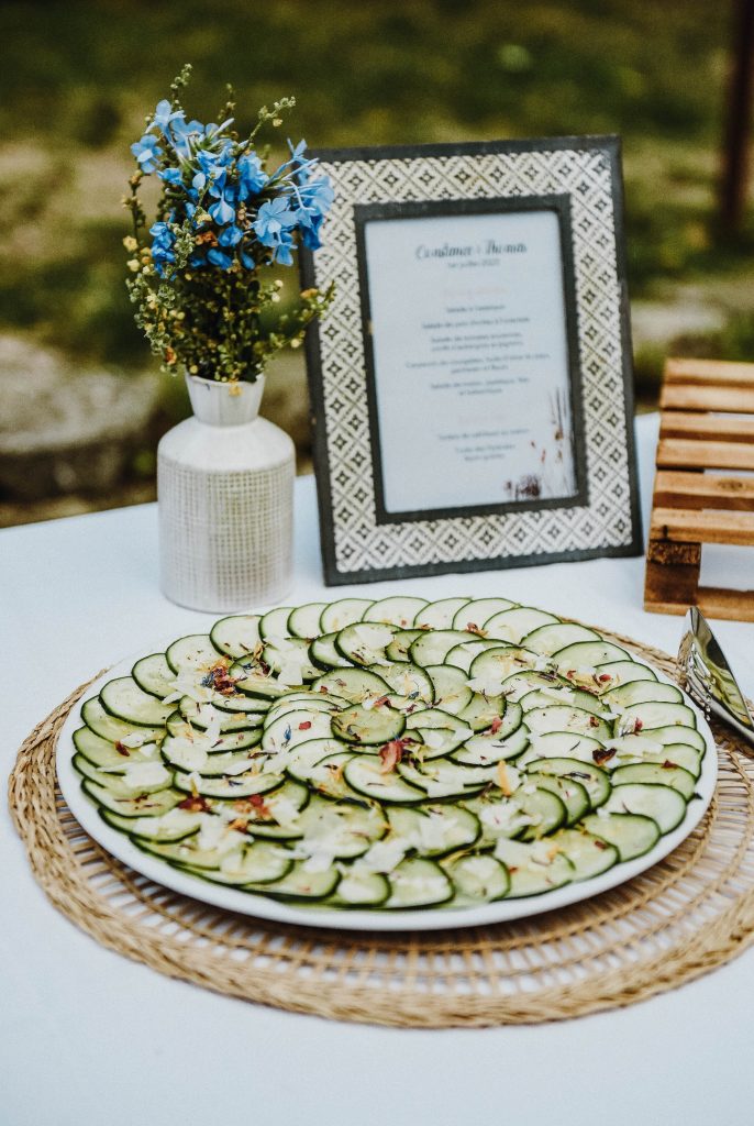 Présentation sur buffet décoré : cadre menu, vase fleuri, set de table naturel, d'un carpaccio de courgettes à l'huile d'olive, fleurs séchées et parmesan.